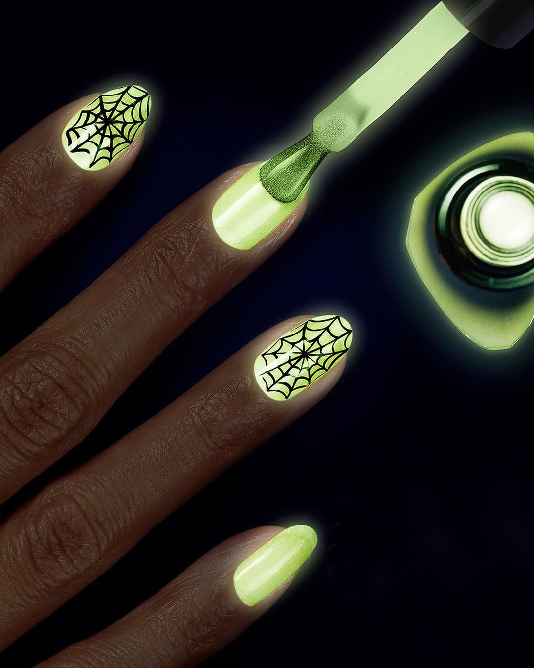 Hand with glowing yellow nails and spiderweb nail art, with nail polish brush hovering above a nail. Shot in dark lighting.