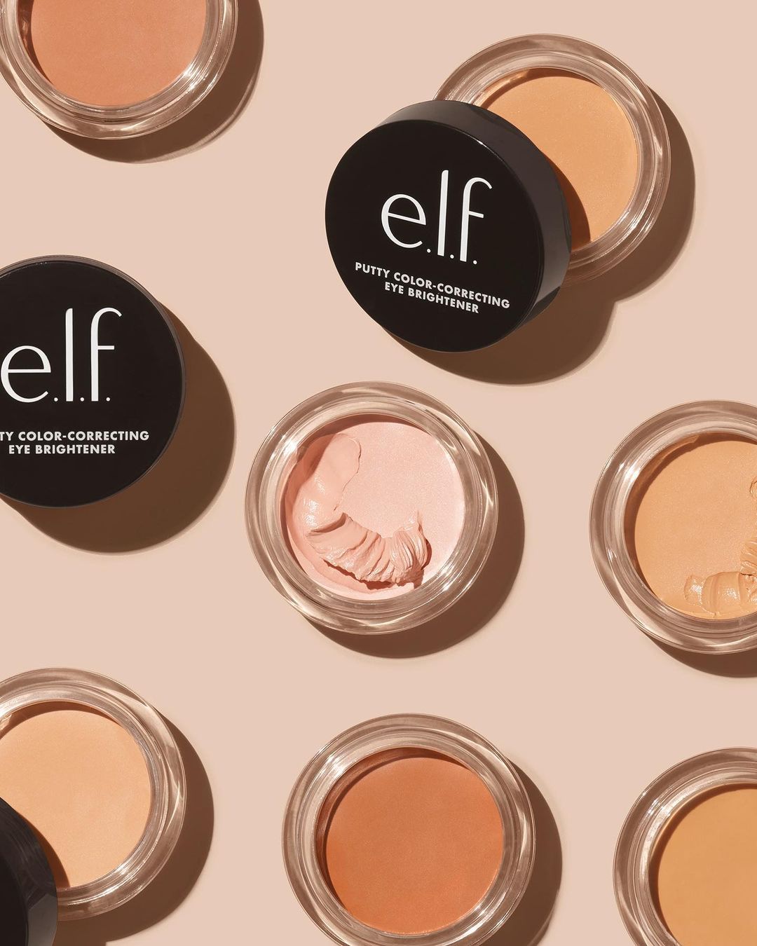 Eye brightener product display with several containers of ELF putty color-correcting cream arranged on a light pink background.
