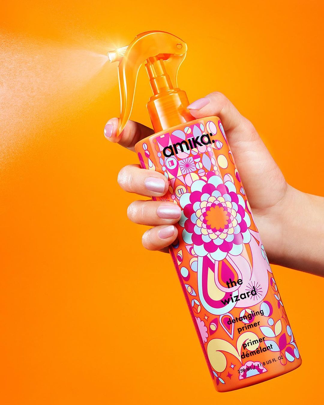 Woman's hand holds Amika The Wizard detangling primer spray bottle with orange nozzle and colorful design against orange background.