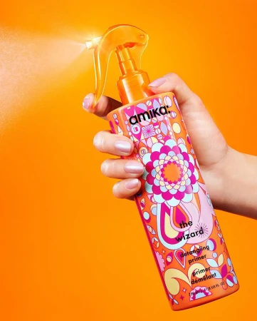 Woman's hand holds Amika The Wizard detangling primer spray bottle with orange nozzle and colorful design against orange background.