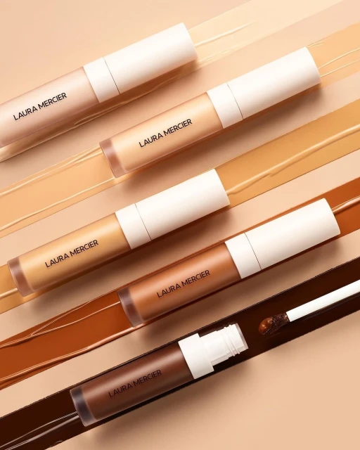 Laura Mercier concealers in varying shades arranged diagonally with corresponding product samples. Beauty and makeup still life.