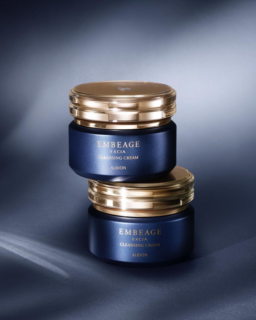 Two dark blue and gold Embeage Excia Cleansing Cream jars stacked against a grey background with soft lighting. Beauty product.