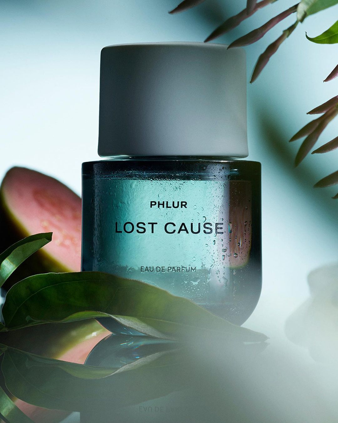 Close-up of a teal perfume bottle with a white cap, featuring the brand name PHLUR and the scent name LOST CAUSE, surrounded by greenery.