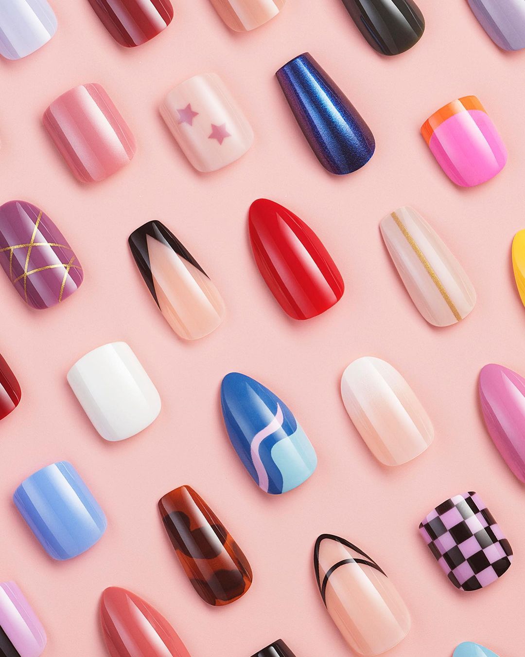 Overhead shot of a colorful collection of press-on nails with varying designs against a solid pink background in a beauty product shot.