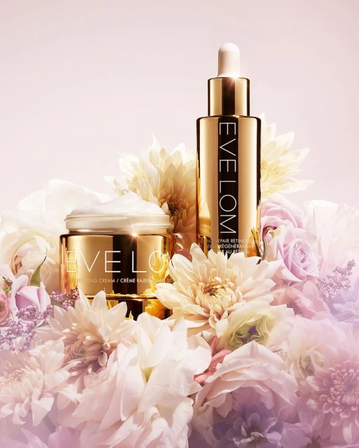 Product shot of Eve Lom skincare products. Retinol elixir and moisturizing cream, featuring gold packaging and white flowers.
