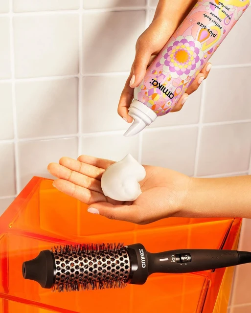 Close-up of hands dispensing Amika hair mousse, alongside the brand's hair brush, set against a textured white and orange backdrop.