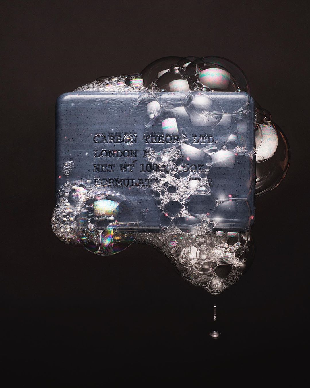 A grey rectangular soap bar with visible text and surrounded by clear soap bubbles against a dark background in a studio setting.