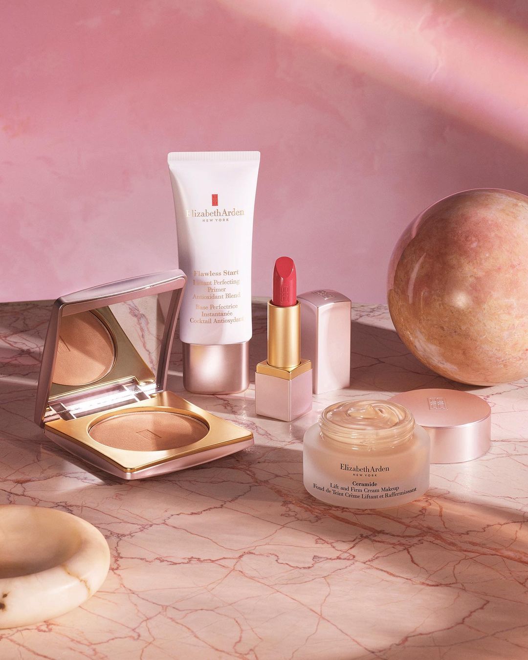 Still life of Elizabeth Arden makeup products including primer, lipstick, bronzer and moisturizer against a pink backdrop.