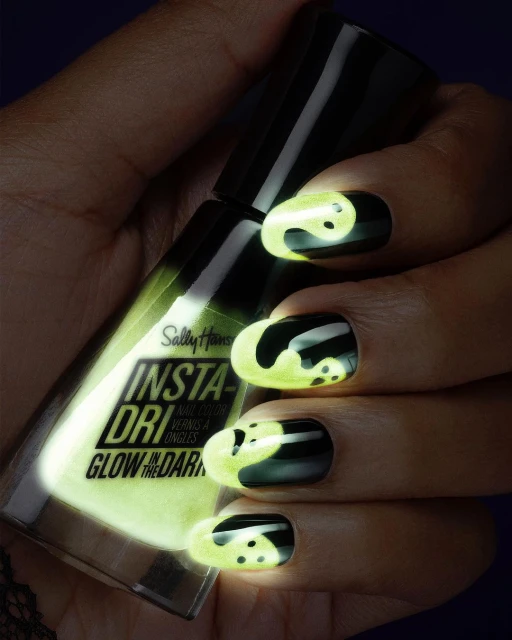 Close-up of a hand holding a bottle of glow-in-the-dark nail polish, with black nails featuring a glowing yin and yang design.