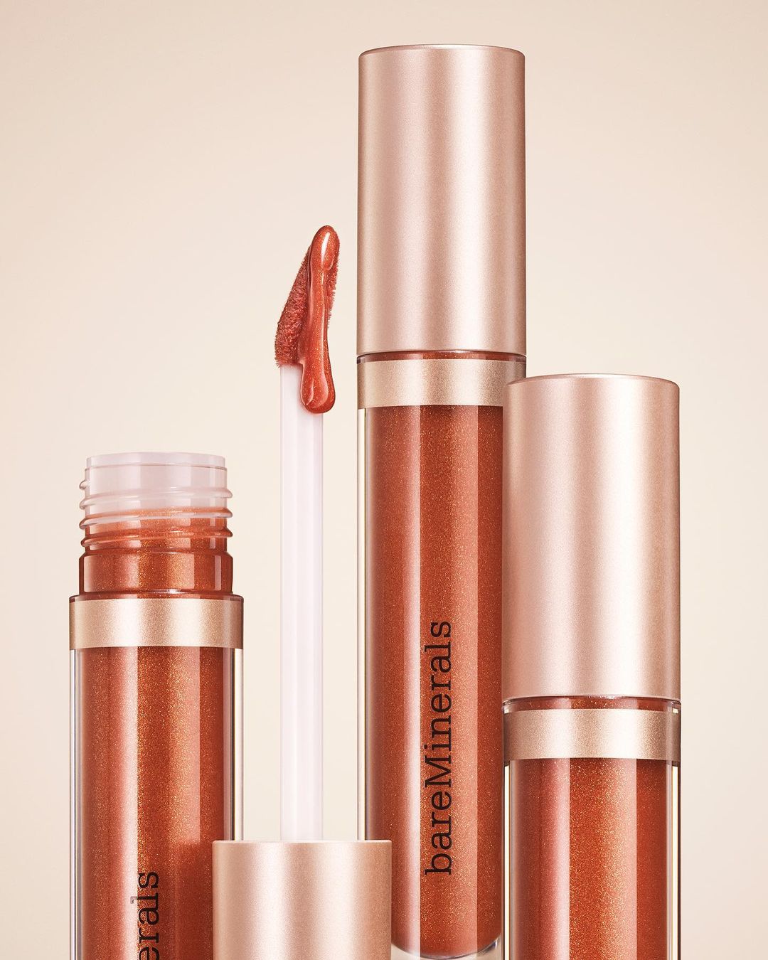 BareMinerals brand lip gloss collection in varying sizes with bronze colored pigment on a beige background. Beauty product photography.