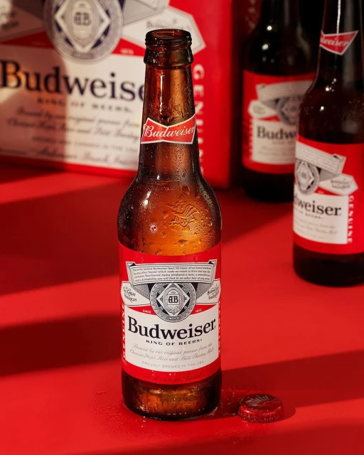 Close-up of a Budweiser beer bottle on a red surface with a bottle cap and other bottles in the soft-focus background.