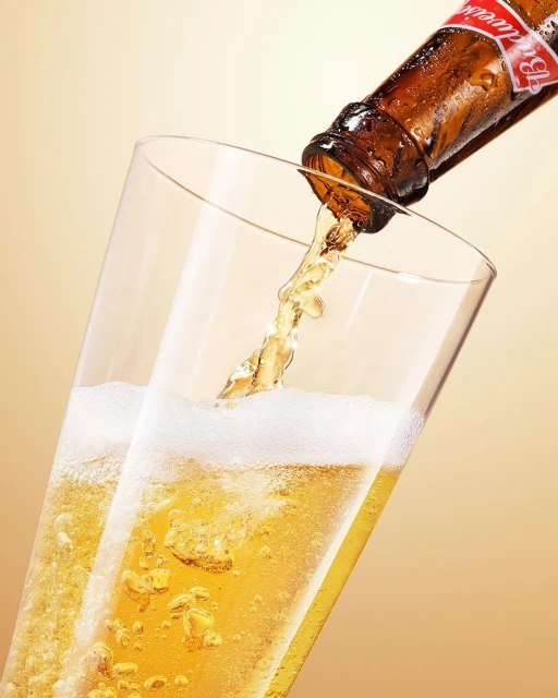 Beer bottle pouring refreshing brew into a tall glass with foamy head against a gradient golden background in a studio setting.