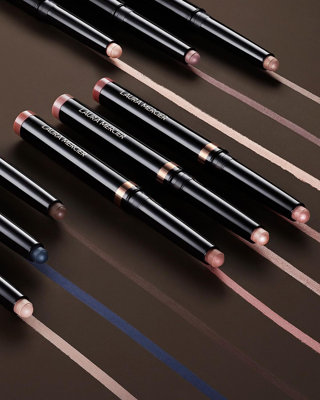 Close-up of several Laura Mercier eye makeup sticks arranged diagonally against a brown backdrop with cosmetic color swatches.