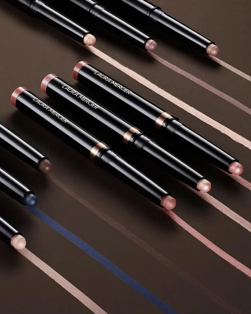 Close-up of several Laura Mercier eye makeup sticks arranged diagonally against a brown backdrop with cosmetic color swatches.