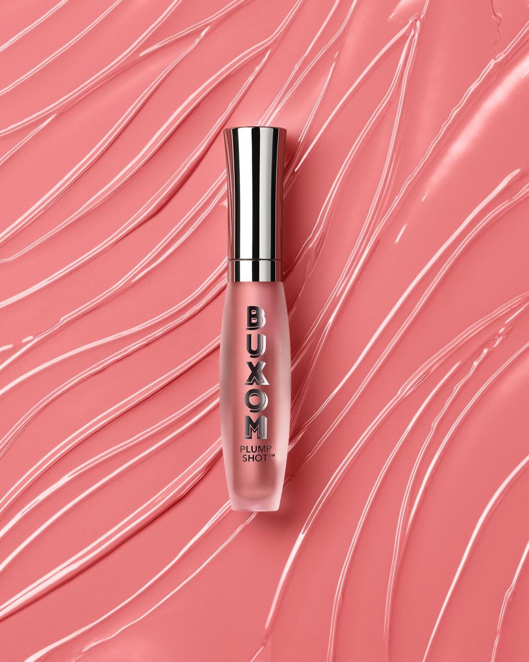 Buxom Plump Shot lip gloss product shot on a peach-colored surface with abstract lines and smears creating a visually dynamic beauty image.