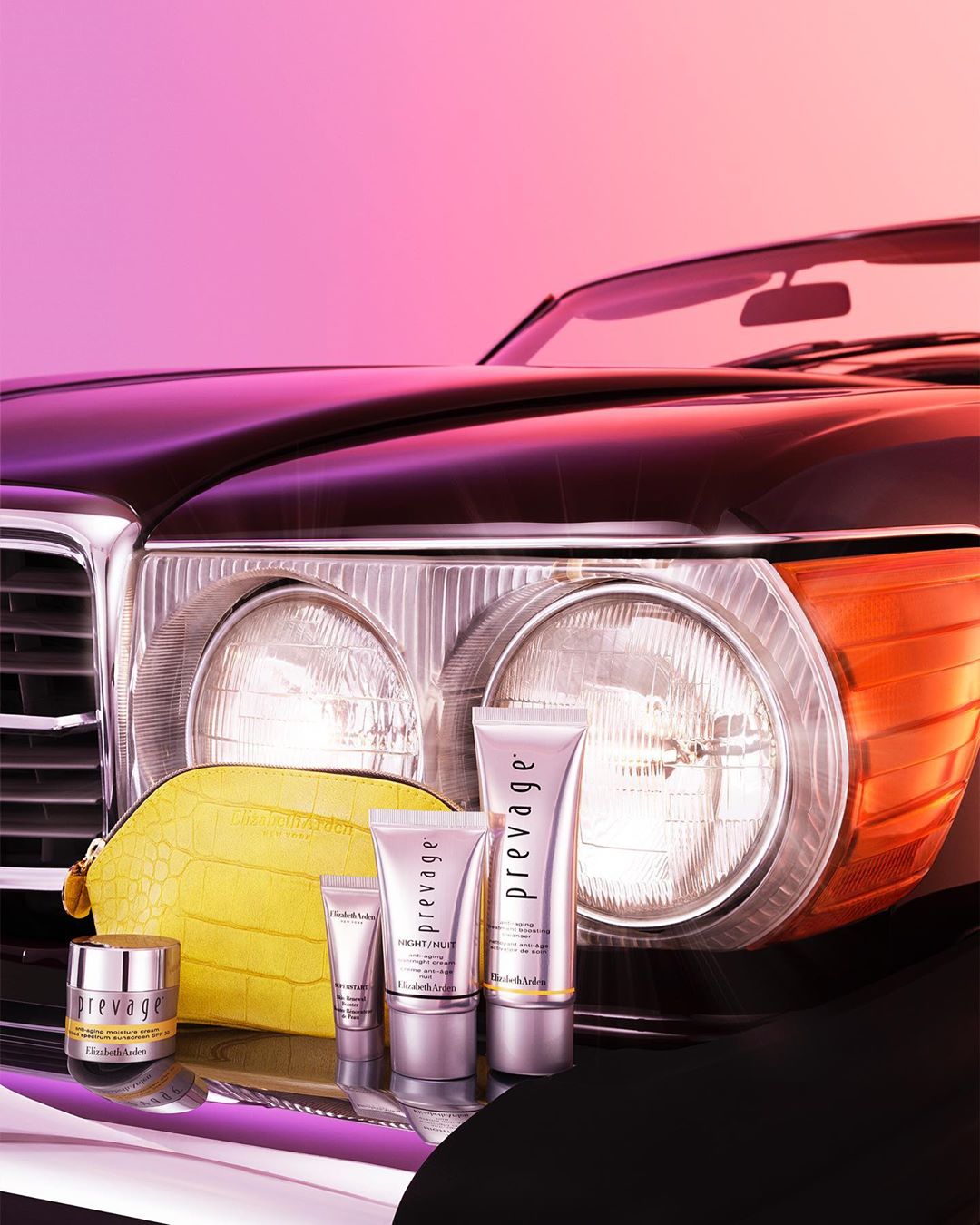 Prevage skincare products on a vintage car hood, featuring cream jars, tubes, and a yellow cosmetic bag against a pink and purple gradient backdrop.