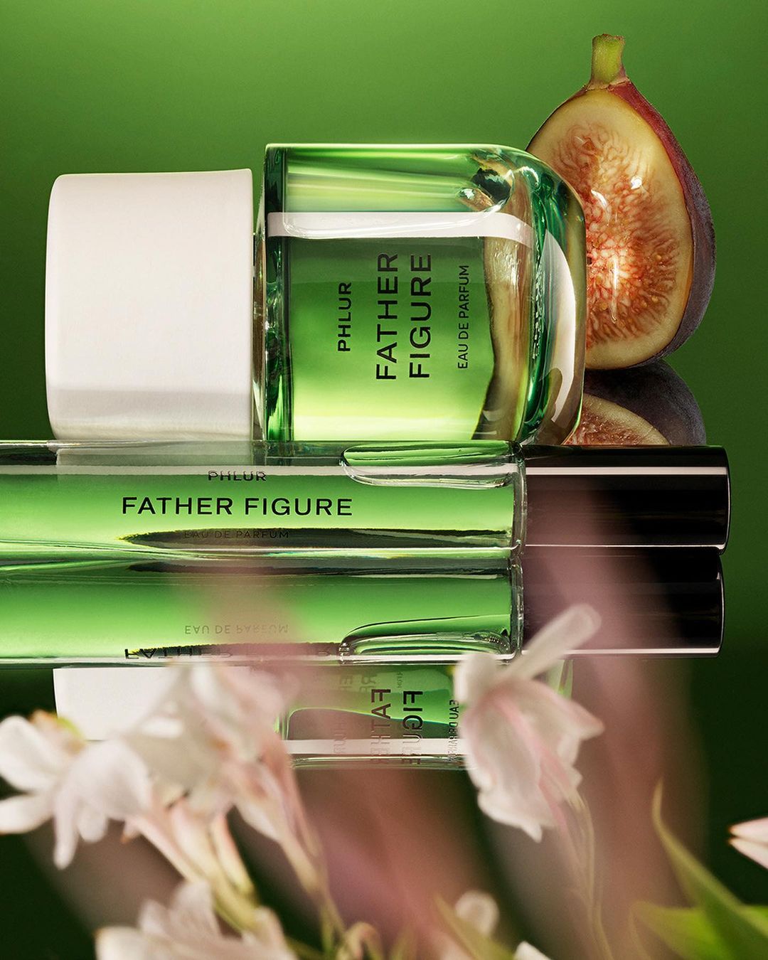Phlur Father Figure perfume bottles displayed on a reflective surface with figs and white flowers against a green background.