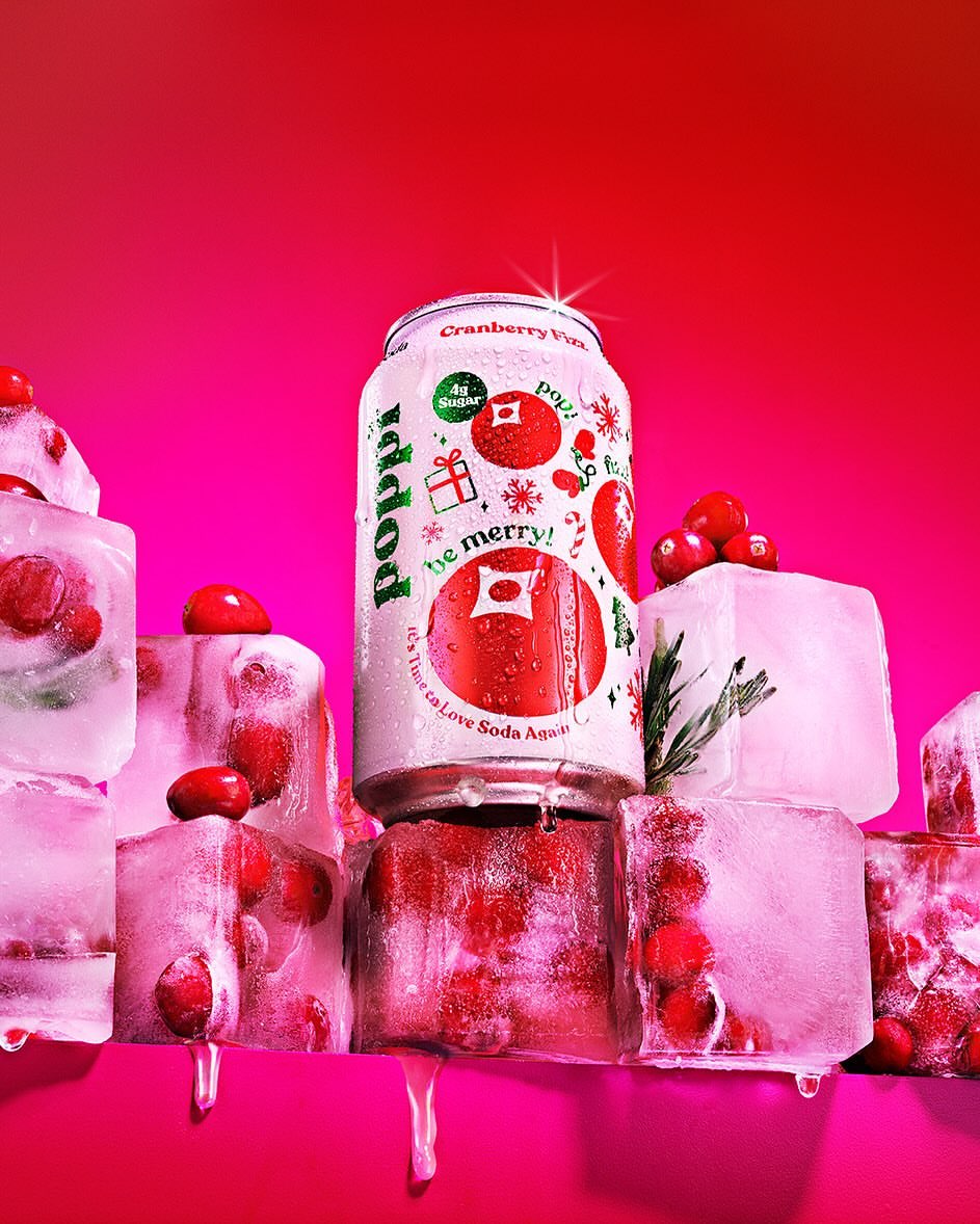 Poppi Cranberry Vibe soda can surrounded by ice cubes and cranberries against a bright pink backdrop in a studio setting.