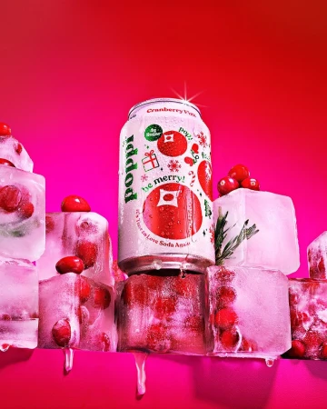 Poppi Cranberry Vibe soda can surrounded by ice cubes and cranberries against a bright pink backdrop in a studio setting.