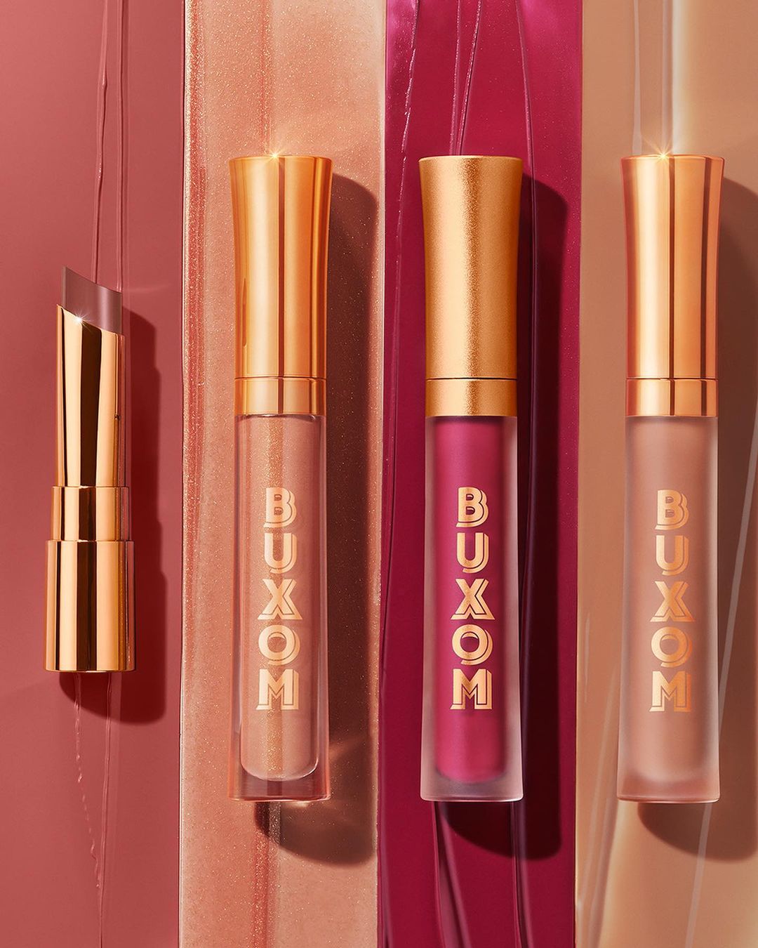 Four Buxom lip products in gold tubes with varying shades of pink and nude, displayed on matching color block surfaces.