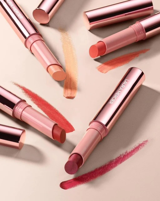 Close-up of Laura Mercier lip and cheek balms in various shades of pink, peach, and red arranged against a neutral background.