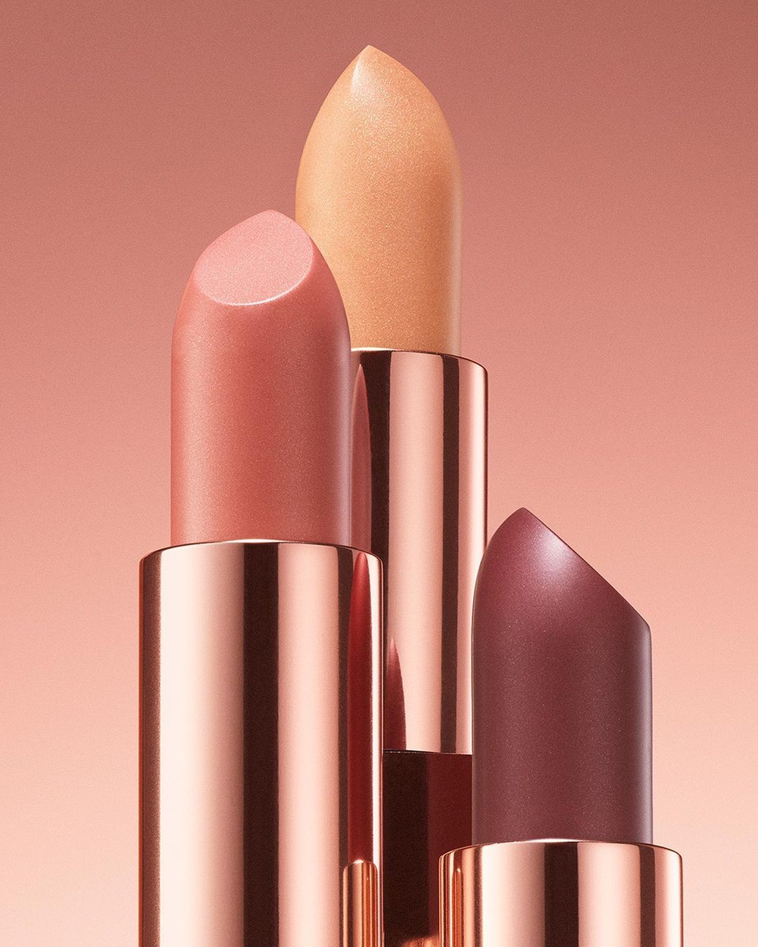 Close-up of three lipsticks in different shades of pink and peach, showcasing cosmetic product photography on a gradient pink background.