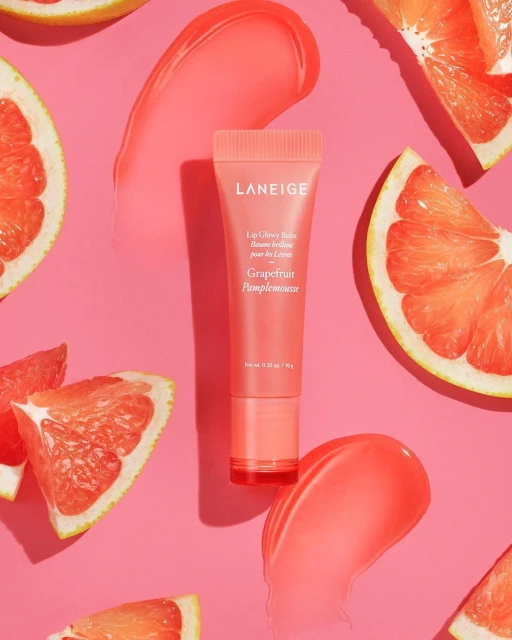 Promotional shot of Laneige lip balm in grapefruit flavor, featuring slices of grapefruit and smears of the lip balm on a pink background.