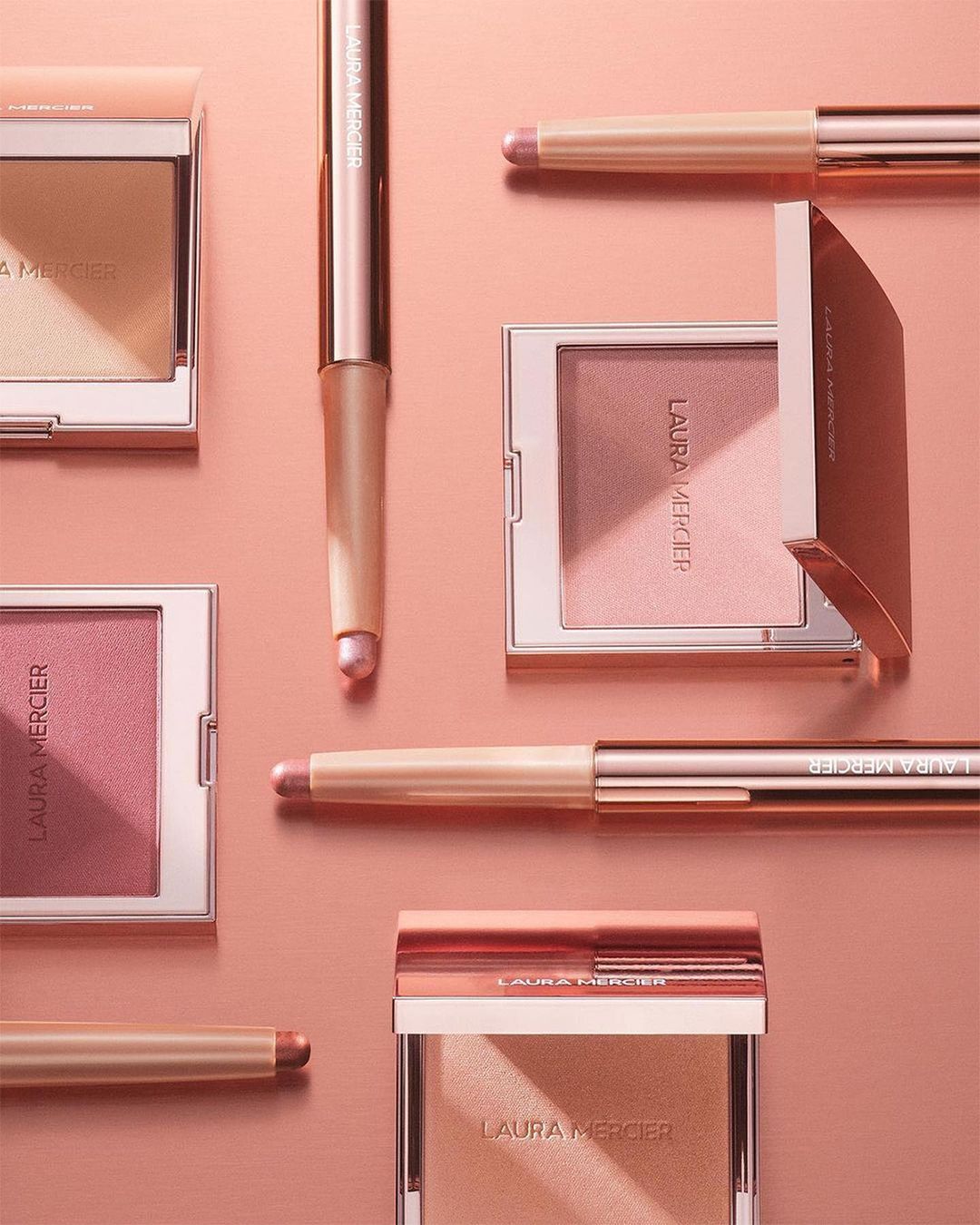Still life of Laura Mercier beauty products including powder compacts and eyeshadow sticks in a pink and cream color palette.