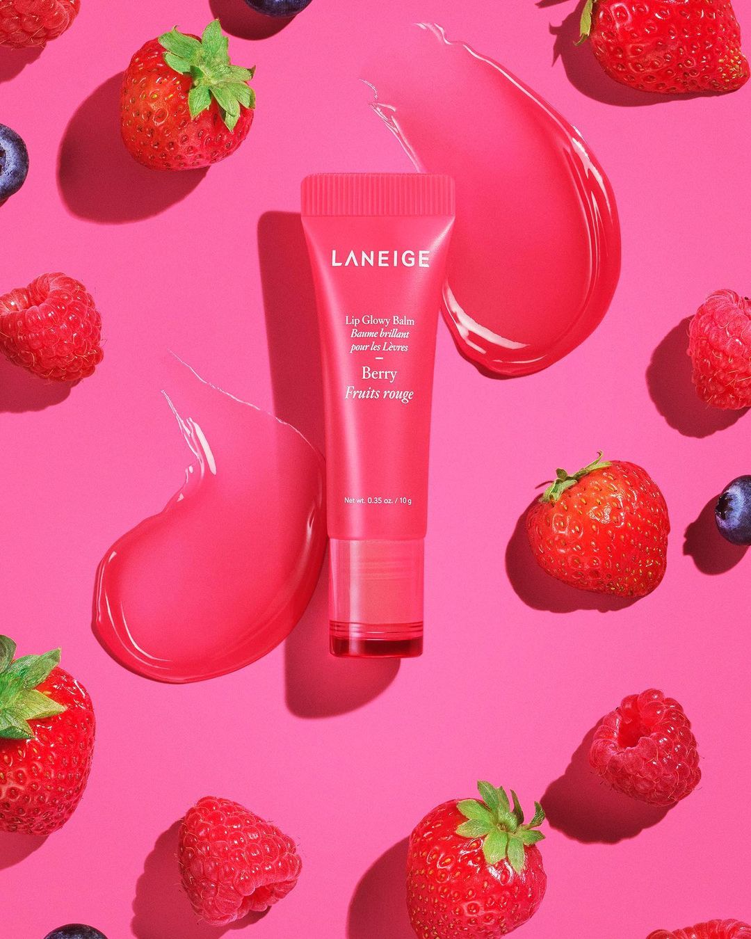Pink tube of Laneige lip gloss surrounded by fresh strawberries, raspberries, and blueberries on a bright pink background. Beauty.