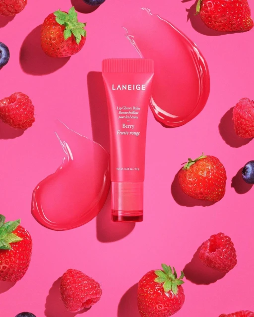 Pink tube of Laneige lip gloss surrounded by fresh strawberries, raspberries, and blueberries on a bright pink background. Beauty.