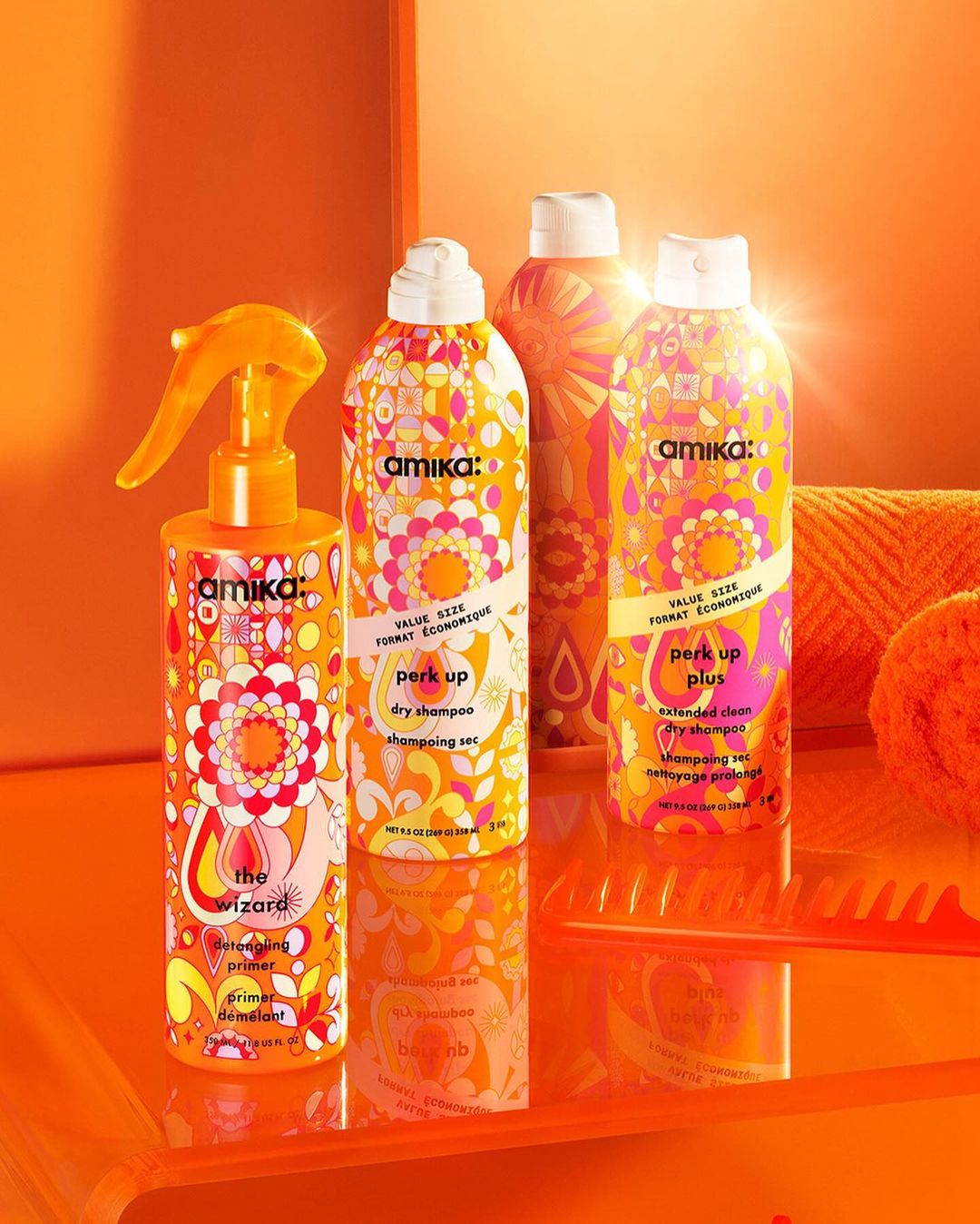 Bright, colorful product shot of Amika hair-care bottles and comb on an orange surface, against a matching orange background.