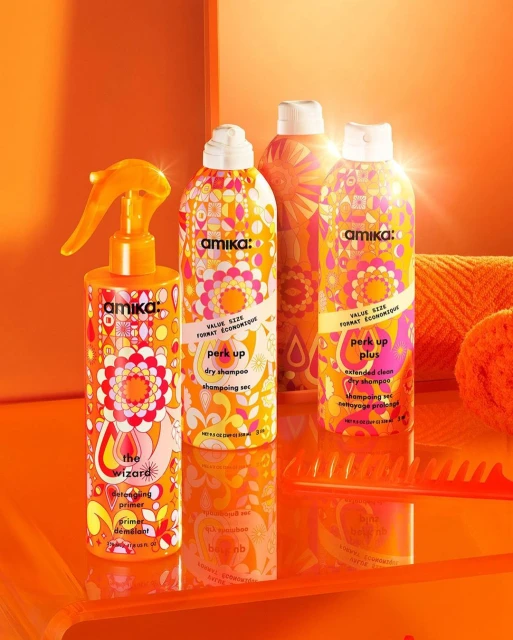 Bright, colorful product shot of Amika hair-care bottles and comb on an orange surface, against a matching orange background.