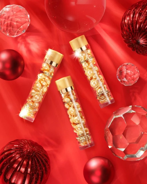 Three Elizabeth Arden Hyaluronic Acid Ceramide Capsules on a red surface surrounded by shiny red decorative spheres. Beauty product.