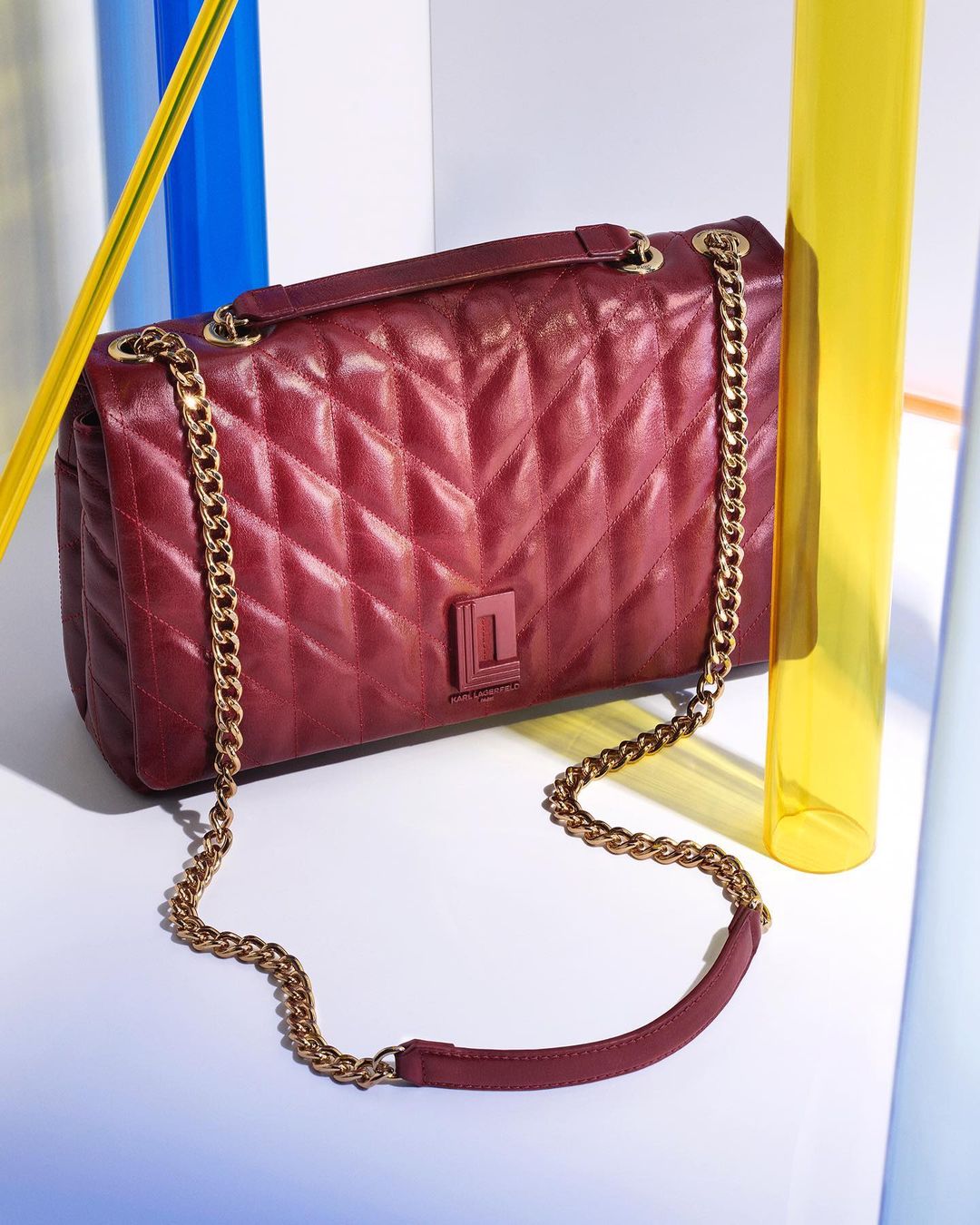 Burgundy quilted leather handbag with gold chain strap and brand logo on a white surface with yellow and blue transparent tubes.