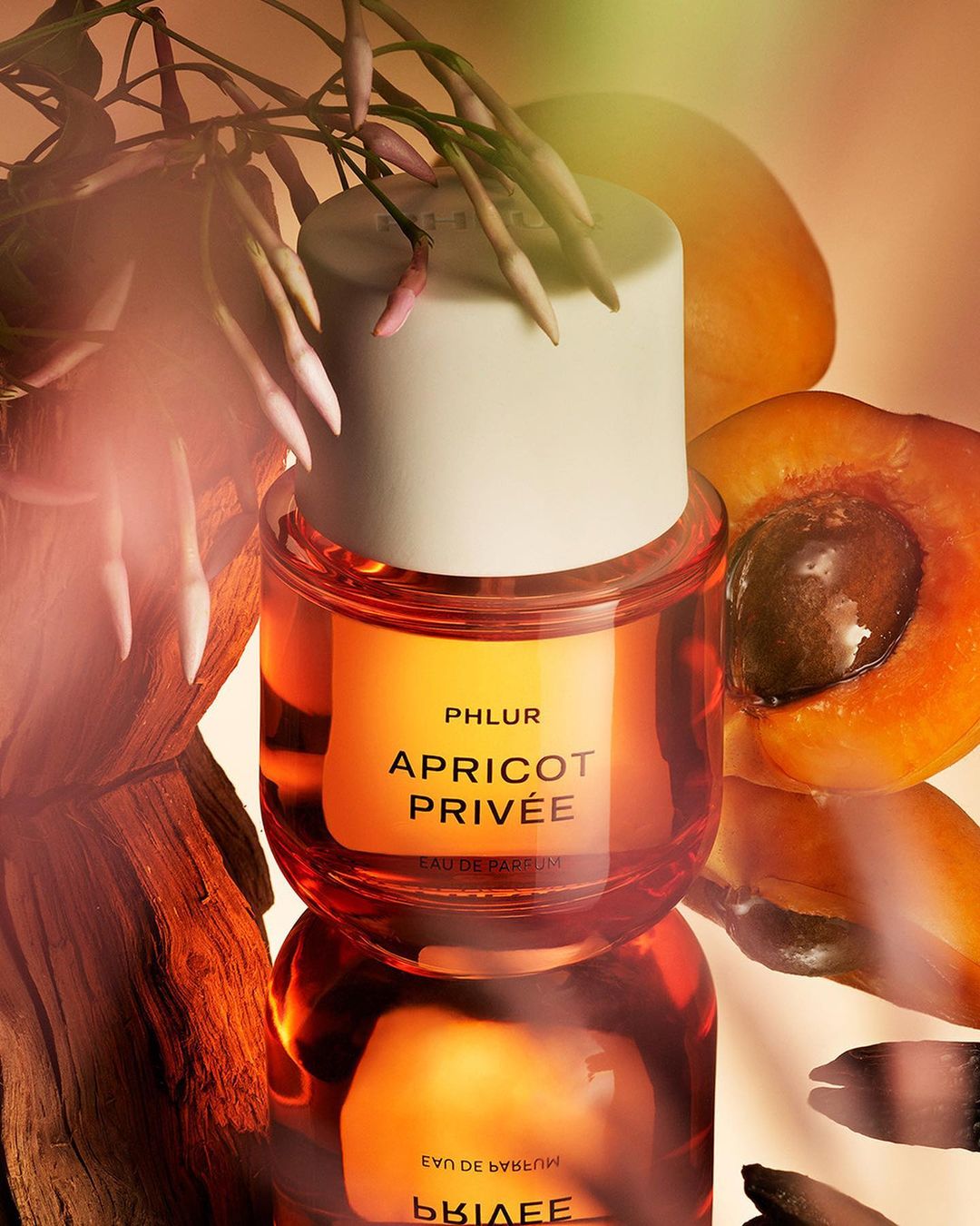 Close-up studio shot of Phlur Apricot Privée perfume bottle with apricot and white flowers, reflecting in glass surface.
