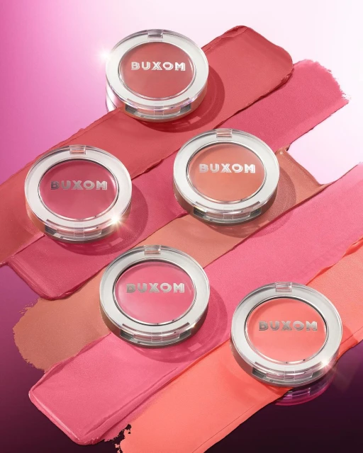 Five Buxom blush compacts are displayed on pink and coral swipes of blush, lit by a soft light and showing off the brand logo.