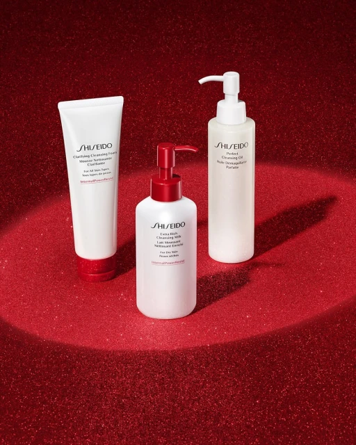 Three Shiseido cleansing products, including a foam, oil, and milk, are arranged on a red glitter surface with spotlight illumination.