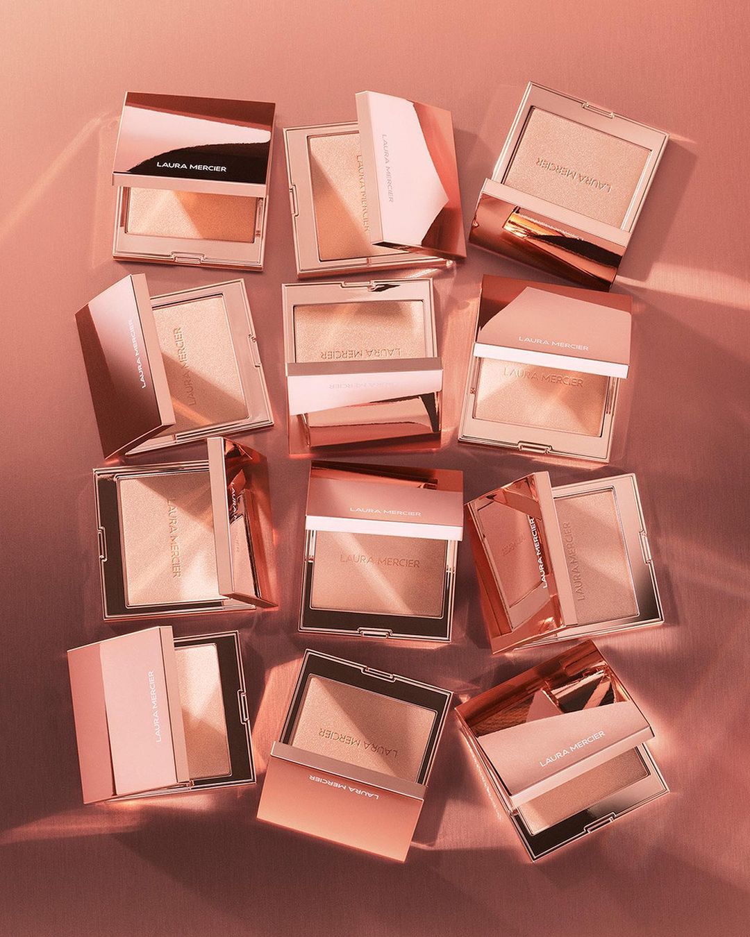 Arrangement of Laura Mercier highlighting powders in mirrored rose gold compacts on a light pink surface, high-angle view, product shot.