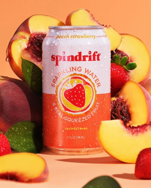 A can of Spindrift sparkling water with peach and strawberry flavors, surrounded by fresh peaches and strawberries, in a bright studio setting.