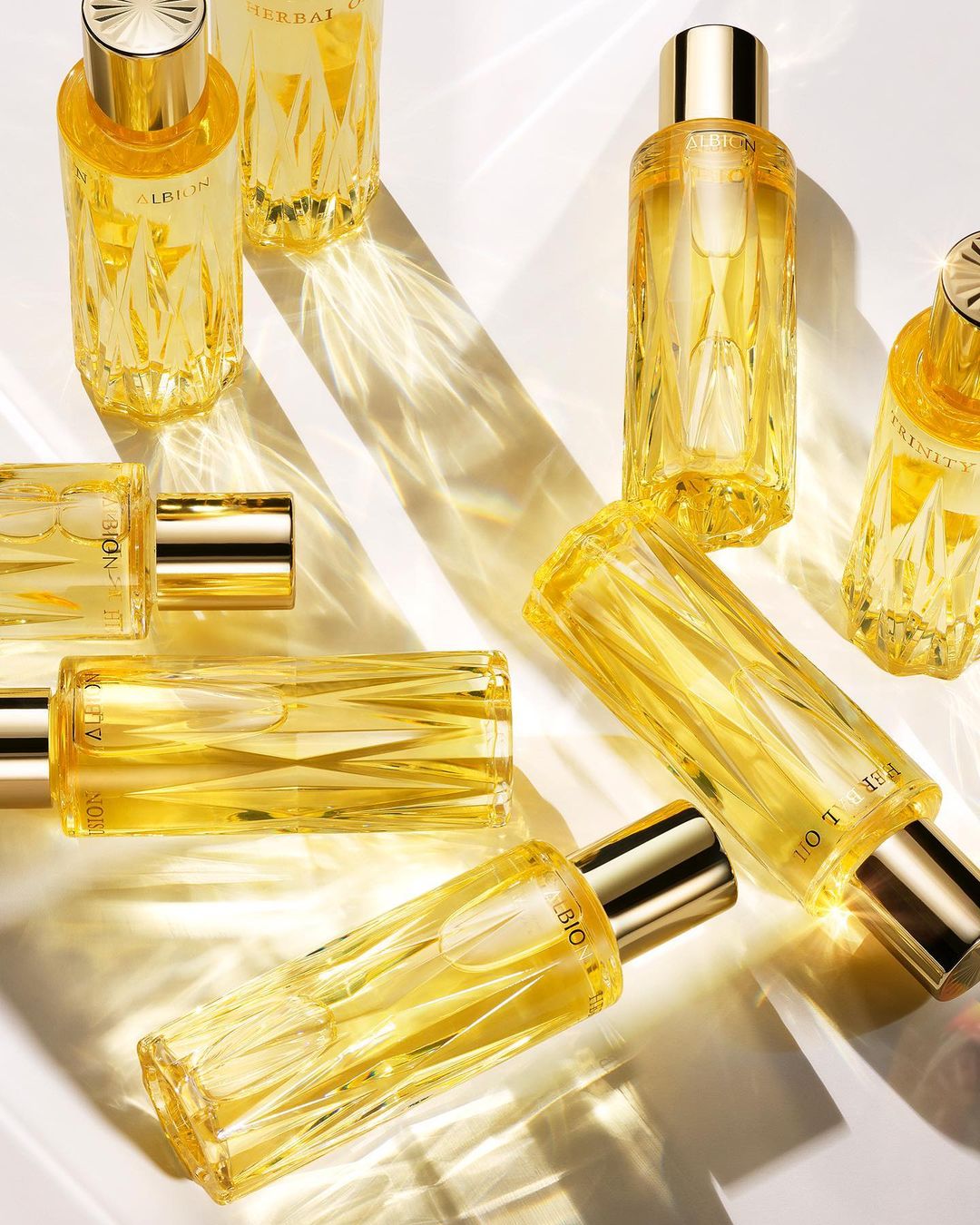 Multiple clear glass bottles with yellow liquid and gold caps are arranged on a reflective surface with light creating abstract patterns.