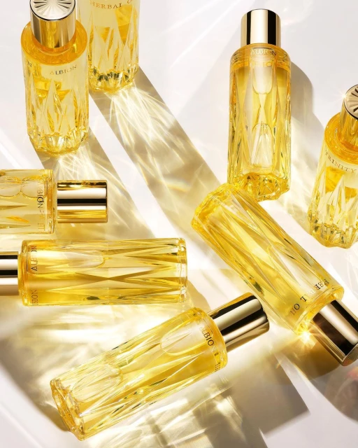 Multiple clear glass bottles with yellow liquid and gold caps are arranged on a reflective surface with light creating abstract patterns.