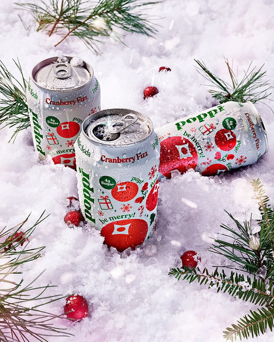 Three cans of Poppi Cranberry Fizz soda are nestled in artificial snow with scattered cranberries and evergreen boughs for a wintry aesthetic.