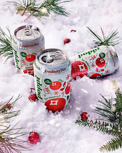 Three cans of Poppi Cranberry Fizz soda are nestled in artificial snow with scattered cranberries and evergreen boughs for a wintry aesthetic.