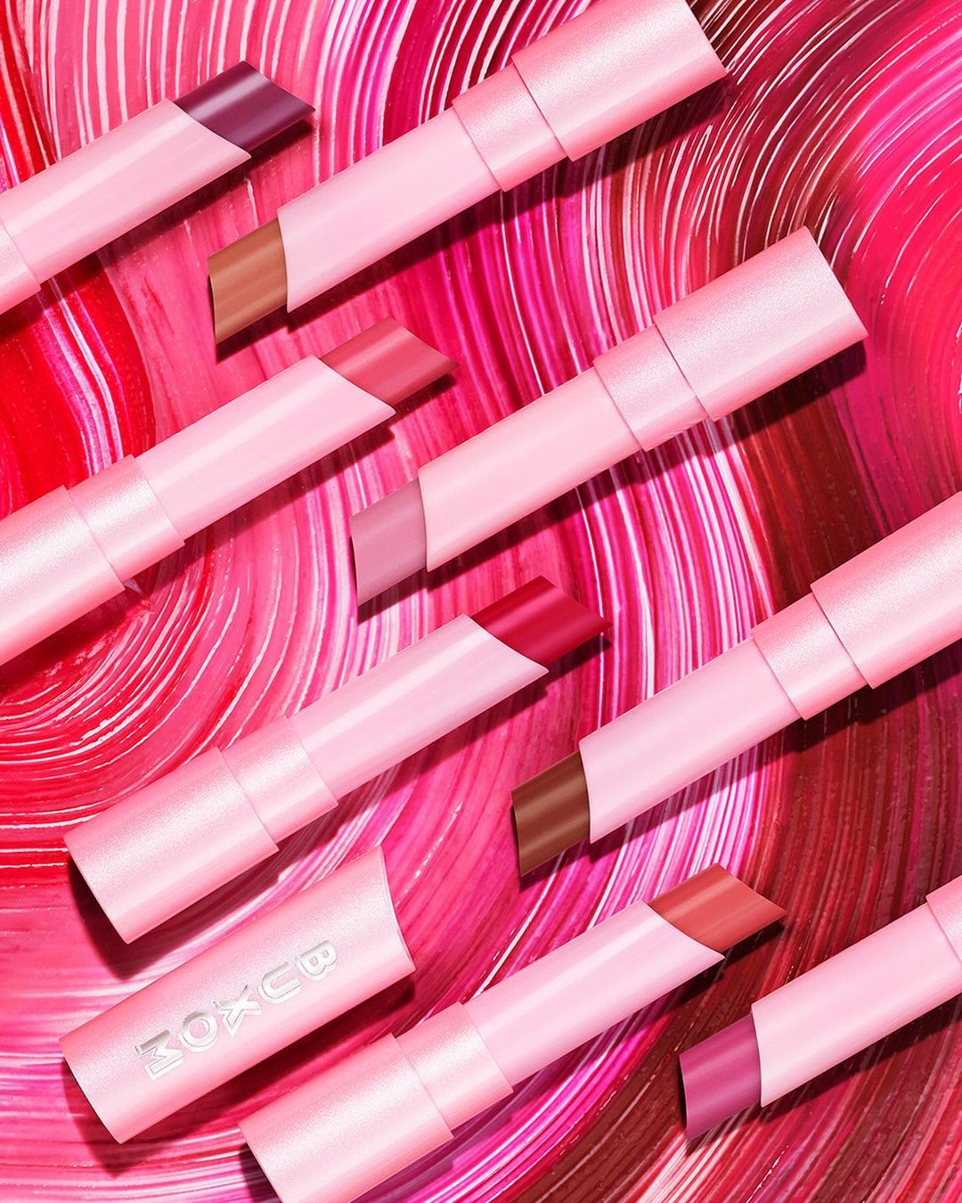 Several pink lipstick tubes with varying shades of color are arranged diagonally against a red and pink swirl background.