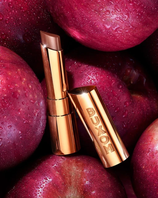 Buxom lipstick in a gold tube with logo, standing among a pile of shiny red apples with water droplets, close-up shot, beauty product