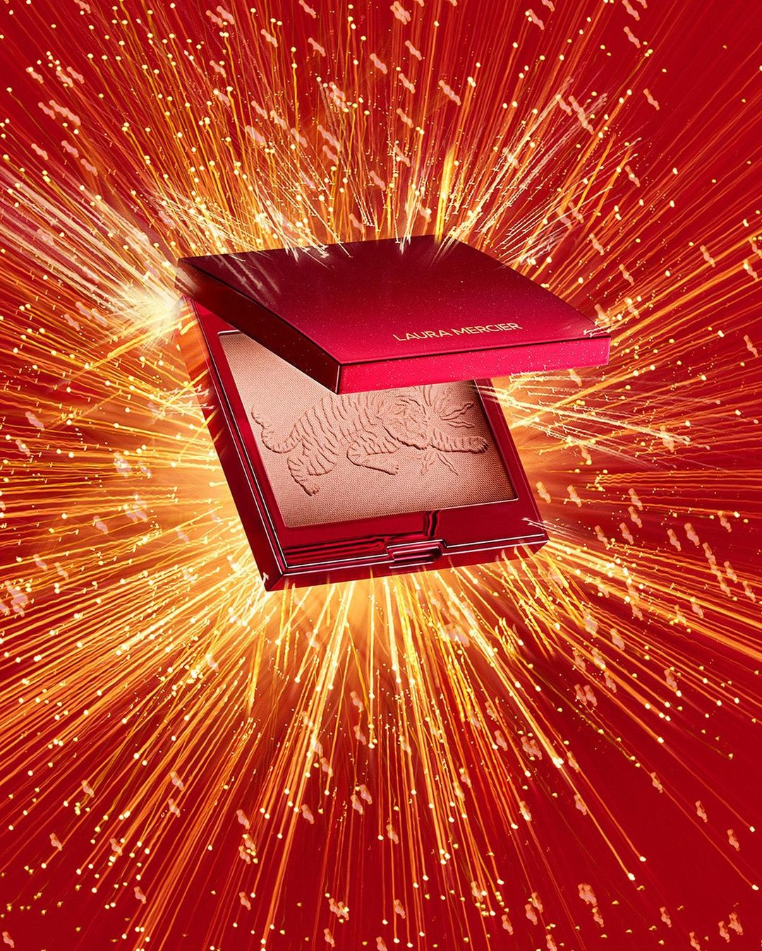 Open red and gold Laura Mercier bronzer with embossed tiger design set against an exploding gold firework background.