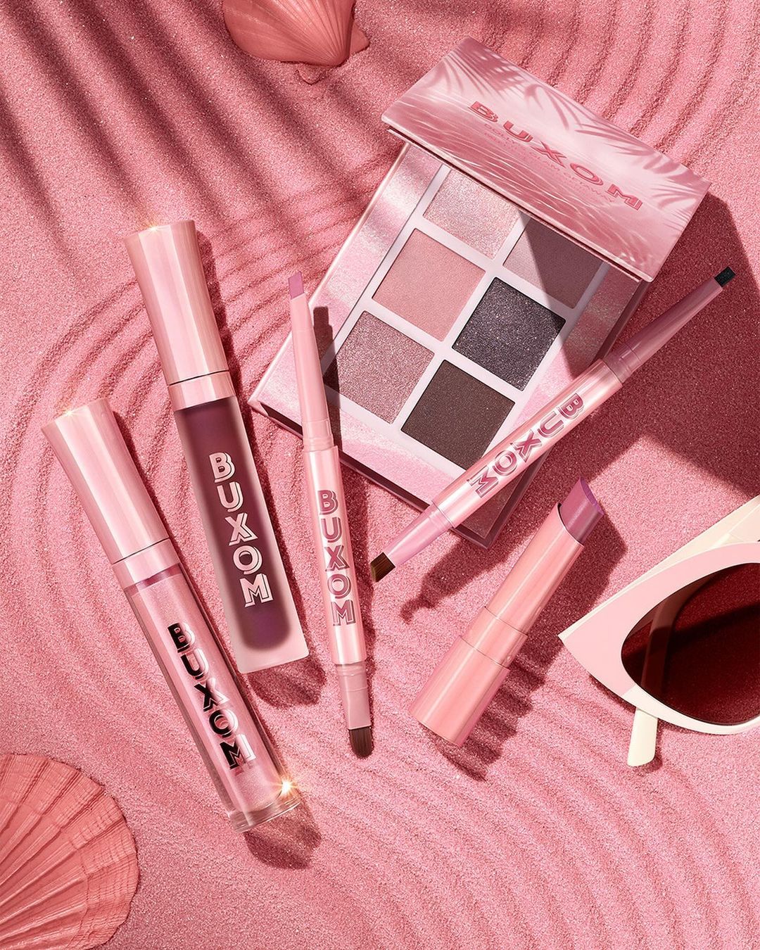 Buxom makeup products arranged on a pink background with soft shadows, including lip gloss, eyeshadow palette, and eyeliner.