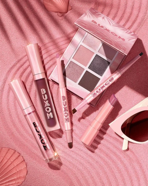 Buxom makeup products arranged on a pink background with soft shadows, including lip gloss, eyeshadow palette, and eyeliner.