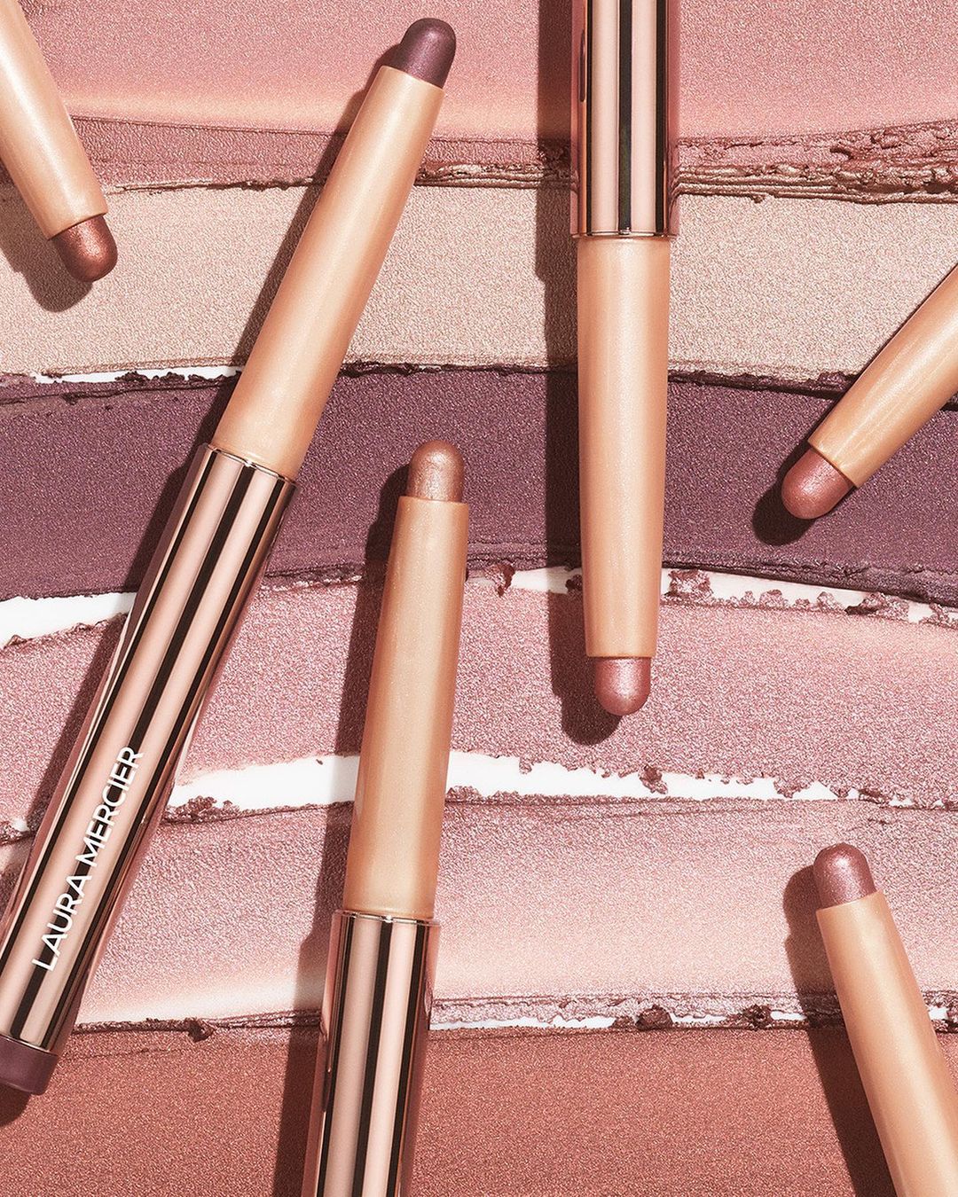 Close-up of Laura Mercier eye shadow sticks in neutral and purple tones on a multi-toned eye shadow palette for a beauty product promotion.