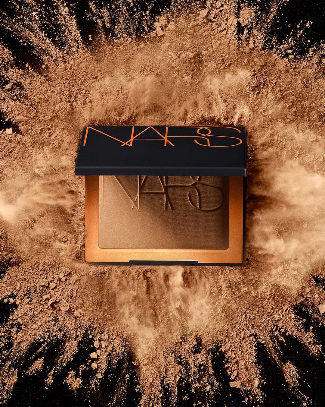NARS bronzing powder compact is displayed against a black background covered in bronzing powder, creating a beauty product shot.